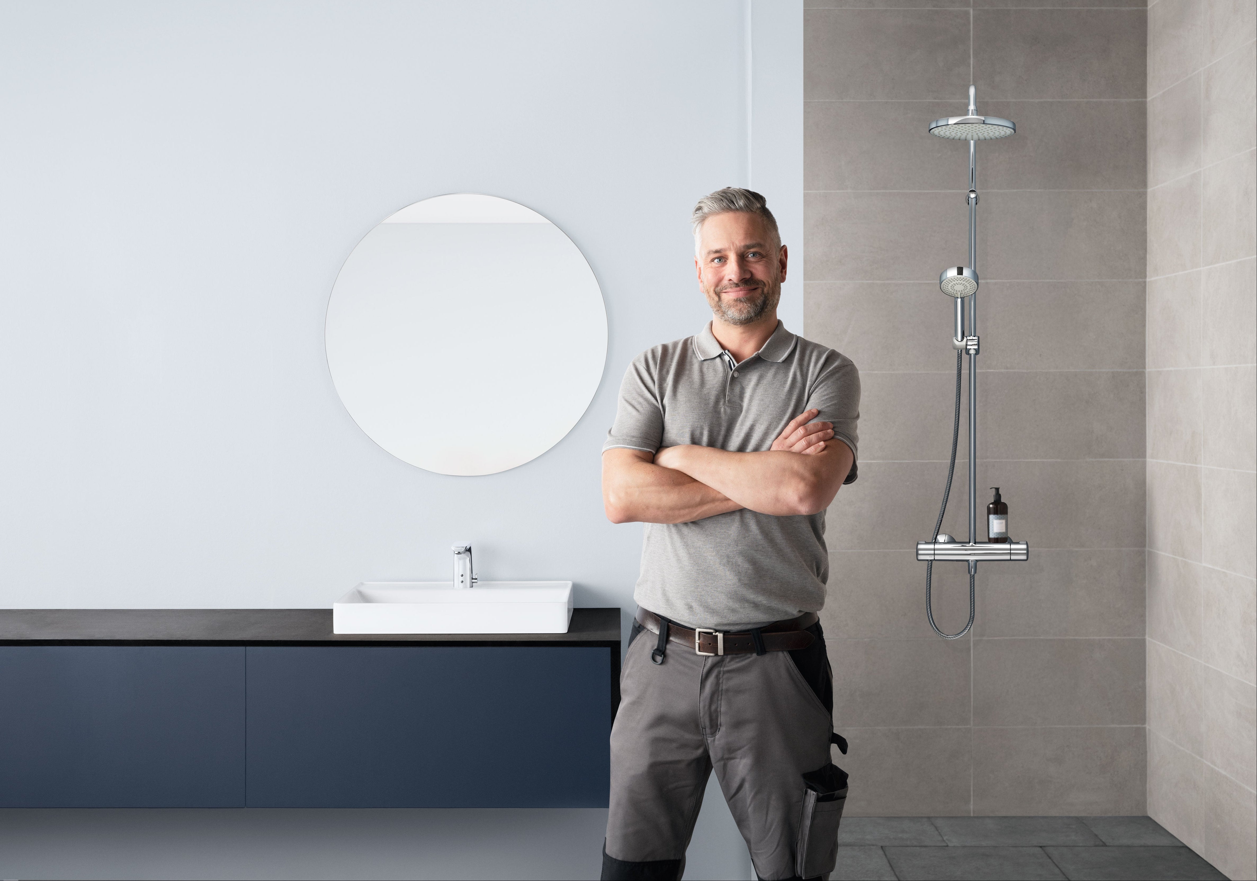 Man standing in a modern bathroom with gray tiles and a shower setup.